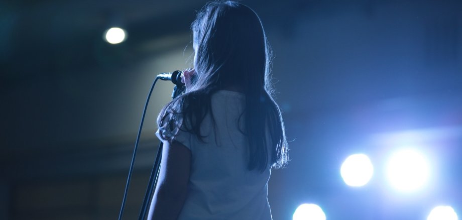 Ein Mädchen steht auf einer Bühne vor einem Mikrofon. Es ist nur von hinten zu sehen. 