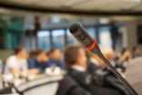 Politik Versammlung Ein Mikrofon im Vordergrund, im unscharfen Hintergrund sitzende Menschen.