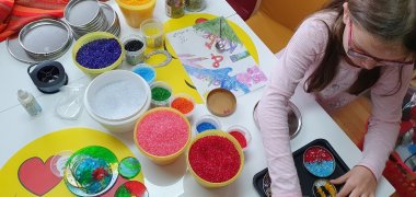 Mitmachmuseum Man sieht ein Mädchen, vor einem Basteltisch, auf dem bunte Glasscheiben und buntes Granulat zum Basteln liegen