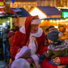Der Weihnachtsmann spricht auf dem Weihnachtsmarkt mit einer Mutter und ihrem Kind, das auf dem Arm gehalten wird.